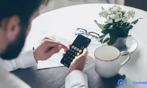 Uomo con barba usa smartphone come calcolatrice per il versamento IVA mensile. Sul tavolo, occhiali, pianta e tazza.
