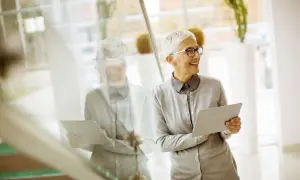 Donna con capelli bianchi e occhiali tiene un tablet. Potrebbe essere un infermiere con partita iva che gestisce la sua attività.