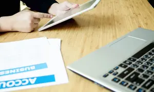 Persona usa un tablet per effettuare un bonifico stessa banca, con documenti e laptop sul tavolo.