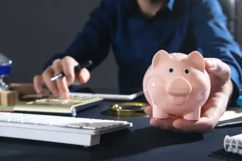 Uomo con salvadanaio rosa calcola come investire i risparmi oggi, con tastiera e calcolatrice a portata di mano.