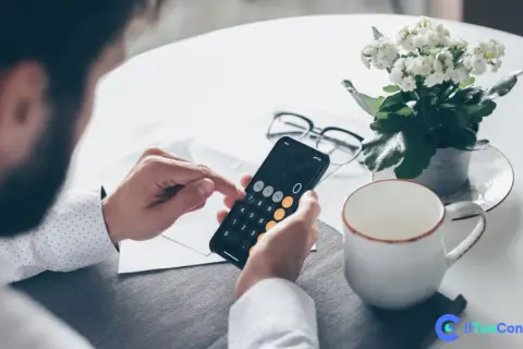 Uomo con barba usa smartphone come calcolatrice per il versamento IVA mensile. Sul tavolo, occhiali, pianta e tazza.