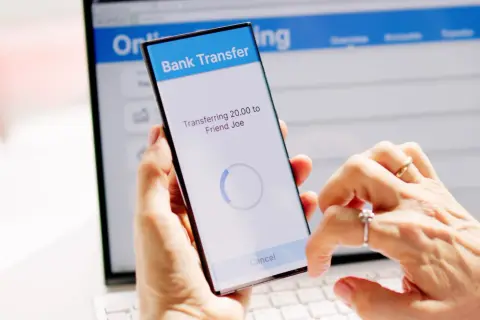 Mani che effettuano un bonifico istantaneo BPER tramite smartphone, con schermo che mostra "Transferring 20.00 to Friend Joe".