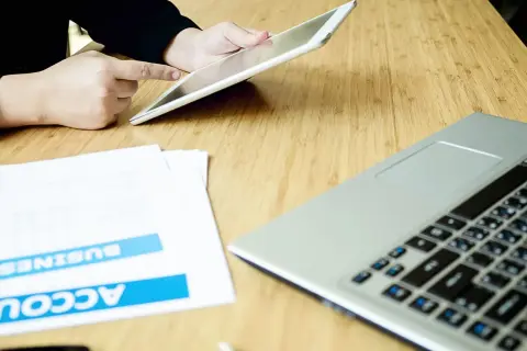 Persona usa un tablet per effettuare un bonifico stessa banca, con documenti e laptop sul tavolo.