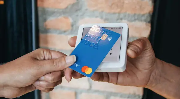 Pagamento contactless con carta blu su POS. La carta mostra "Soma com você" e i loghi Mastercard e Visa.