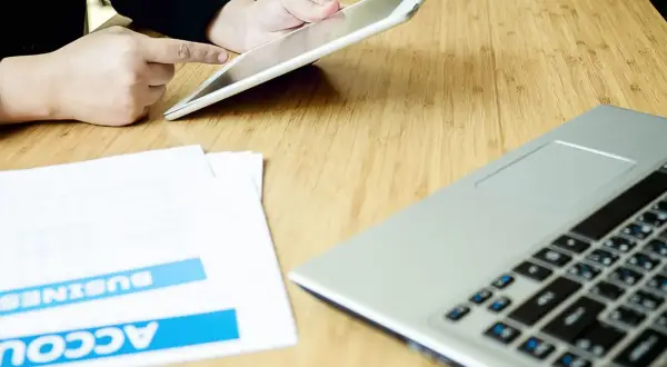 Persona usa un tablet per effettuare un bonifico stessa banca, con documenti e laptop sul tavolo.