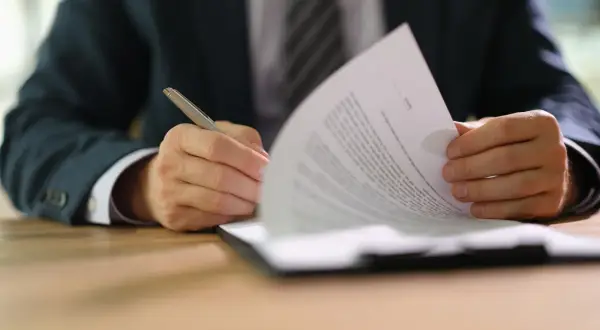 Uomo in giacca e cravatta firma un documento, parte di un set informativo.
