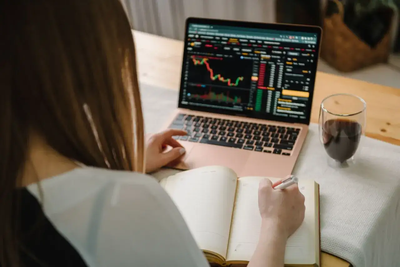Donna studia grafici finanziari sul laptop, prendendo appunti per il suo trading online. Un caffè la accompagna.