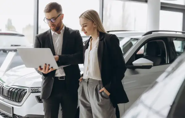 Uomo e donna in un concessionario auto esaminano le specifiche di una nuova macchina aziendale su un laptop.