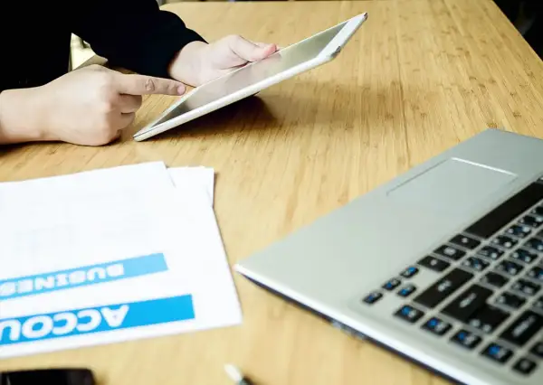 Persona usa un tablet per effettuare un bonifico stessa banca, con documenti e laptop sul tavolo.